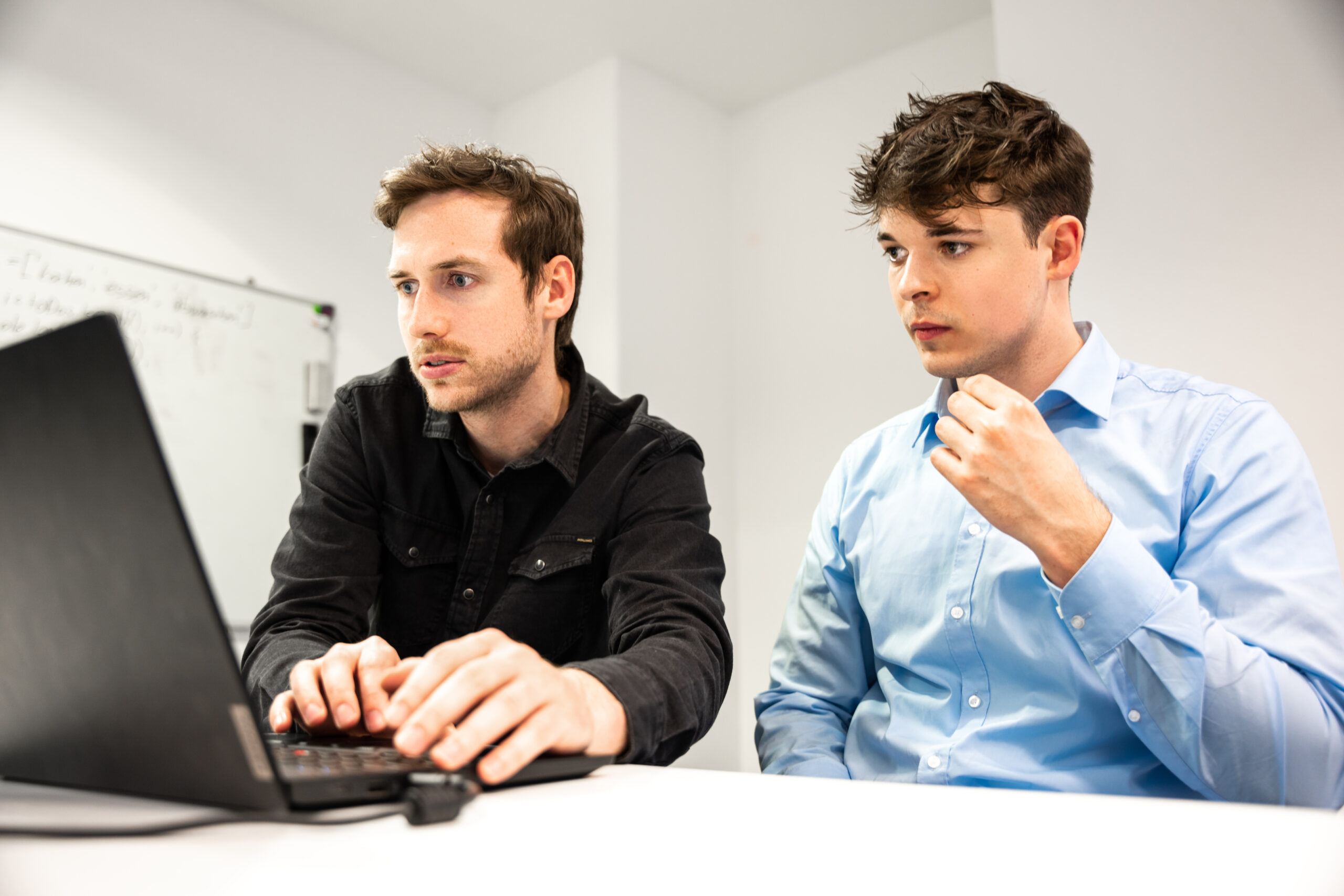 Zwei Männer sitzen an einem Tisch vor einem Laptop. Die linke Person bedient den Laptop. Die rechte Person schaut gespannt zu.