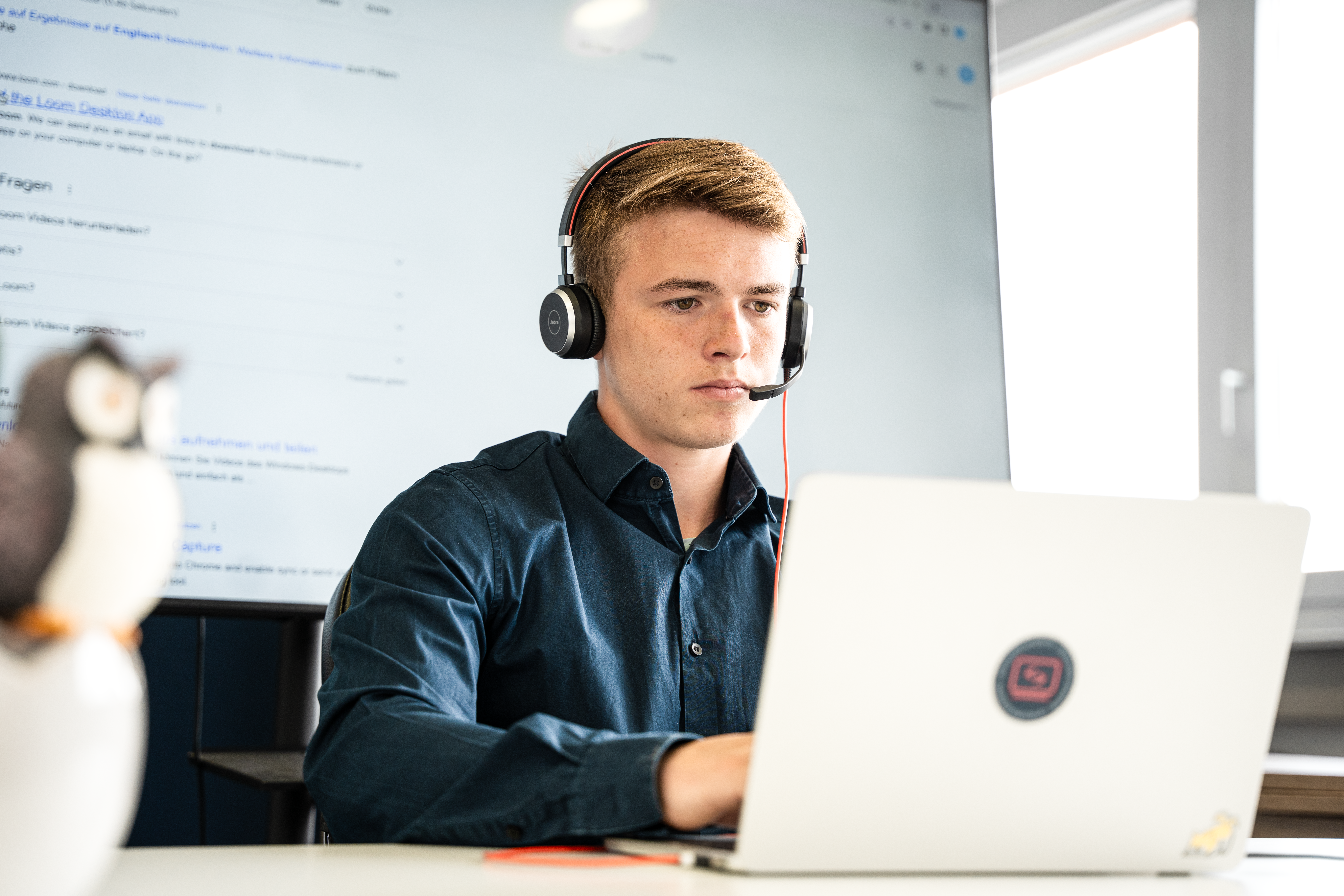 Person mit Kopfhörern sitzt an einem Tisch vor einem Laptop und codet. Im Hintergrund ist ein großer Bildschirm mit einem Internet-Suchverlauf.