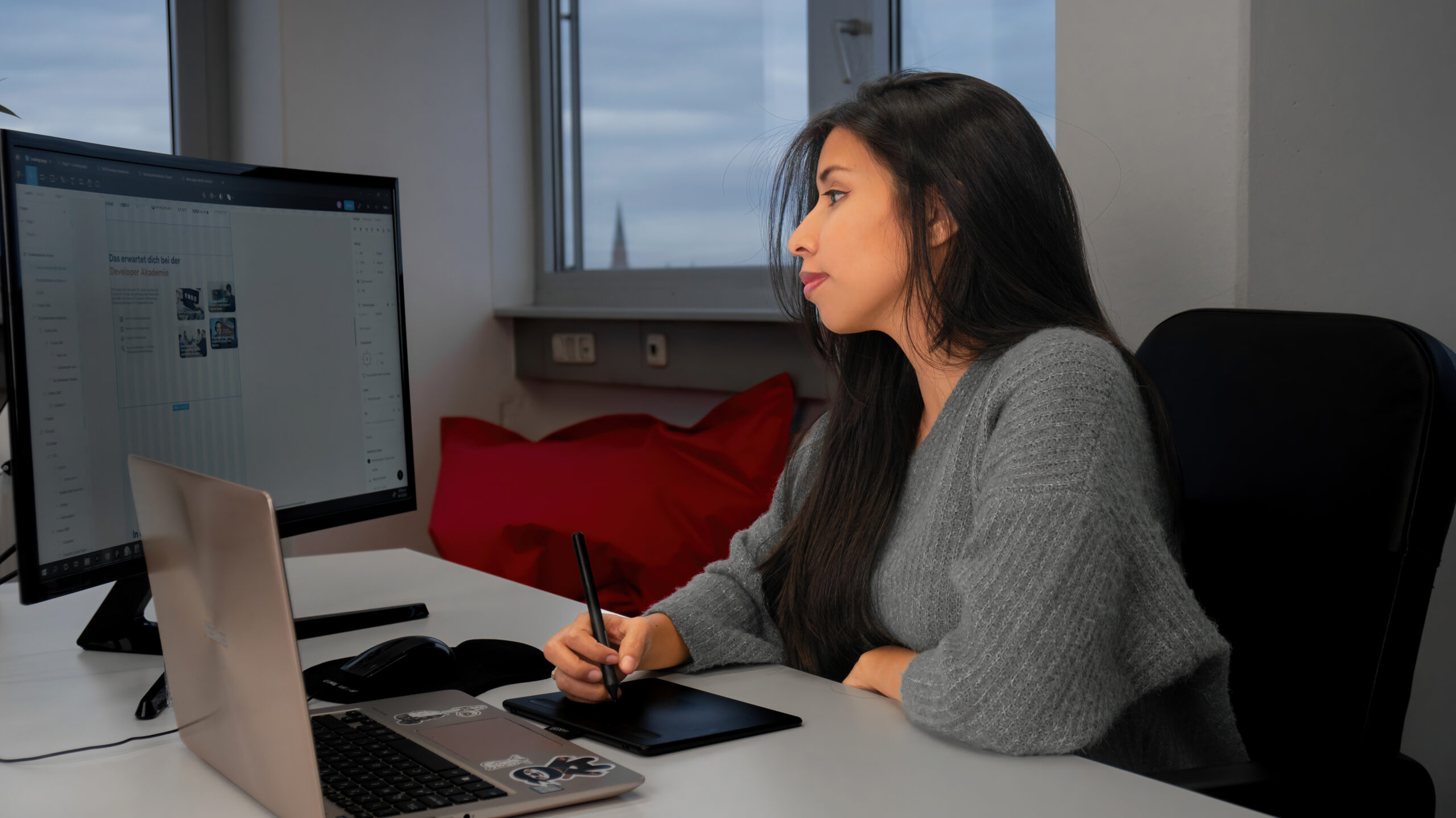 Frau sitzt an Schreibtisch mit Laptop und Computerbildschirm. Sie schaut auf den Bildschirm und designt eine Webseite.